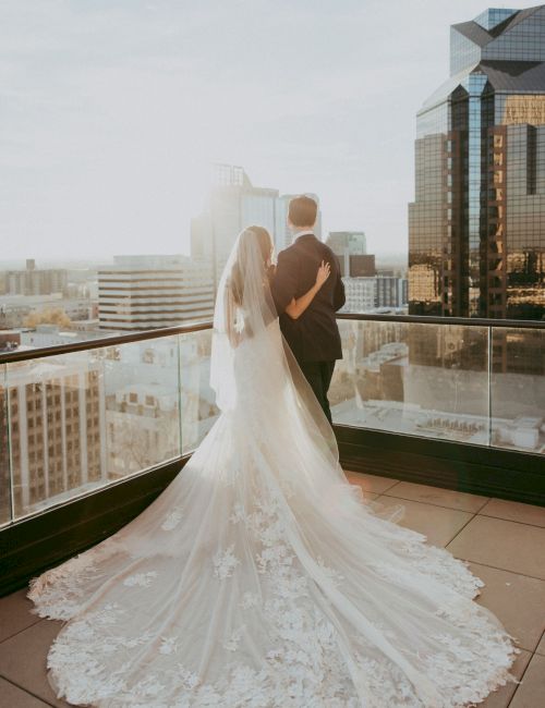 A bride and groom in wedding attire stand on a high-rise terrace, embracing with a city skyline behind them.