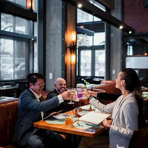 Three people in a modern restaurant toast with glasses, smiling and celebrating together over drinks and dinner.